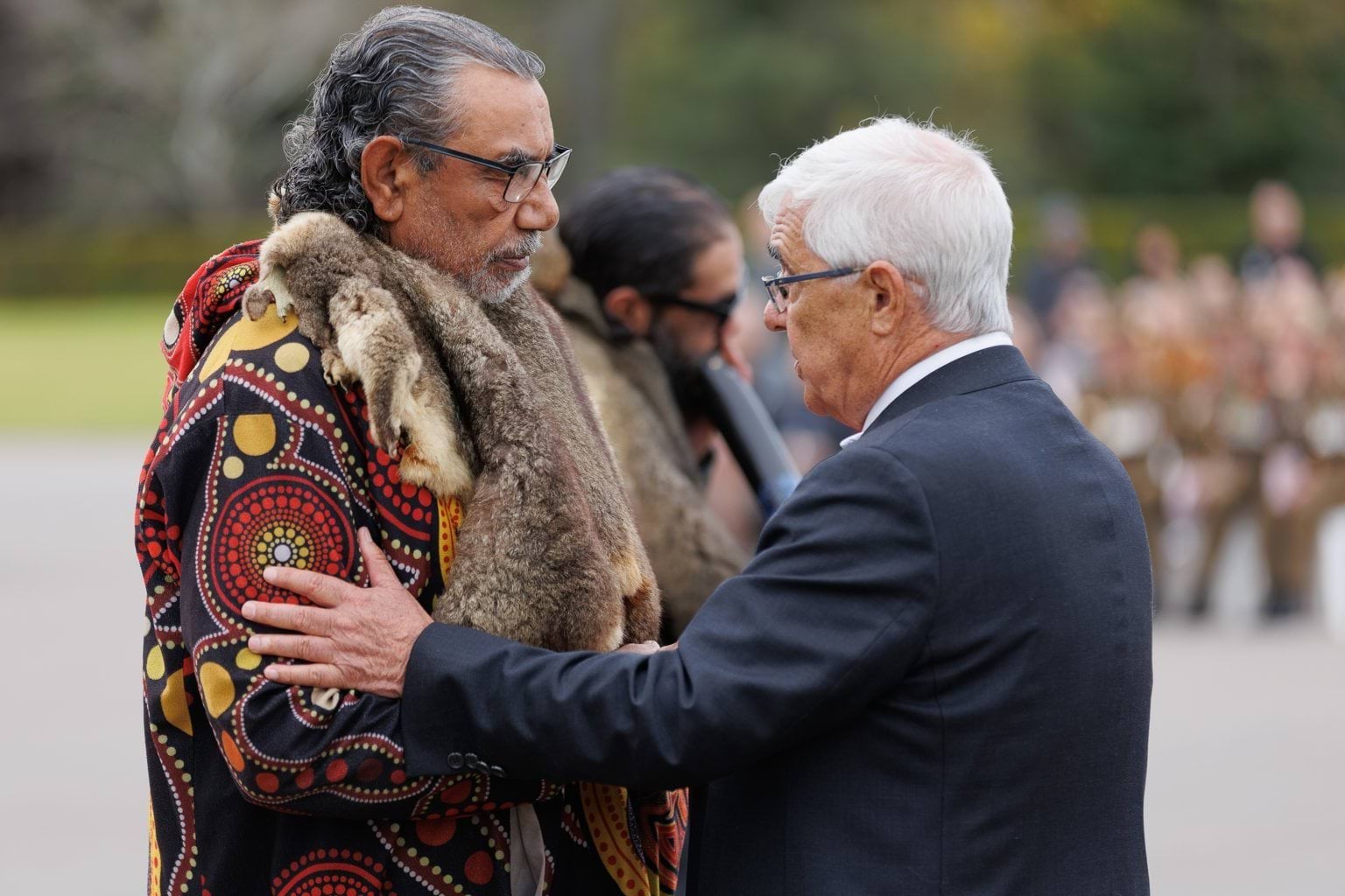 Uncle Charles Pakana and Uncle Mick Edwards embrace as a man plays the didgeridoo in the background