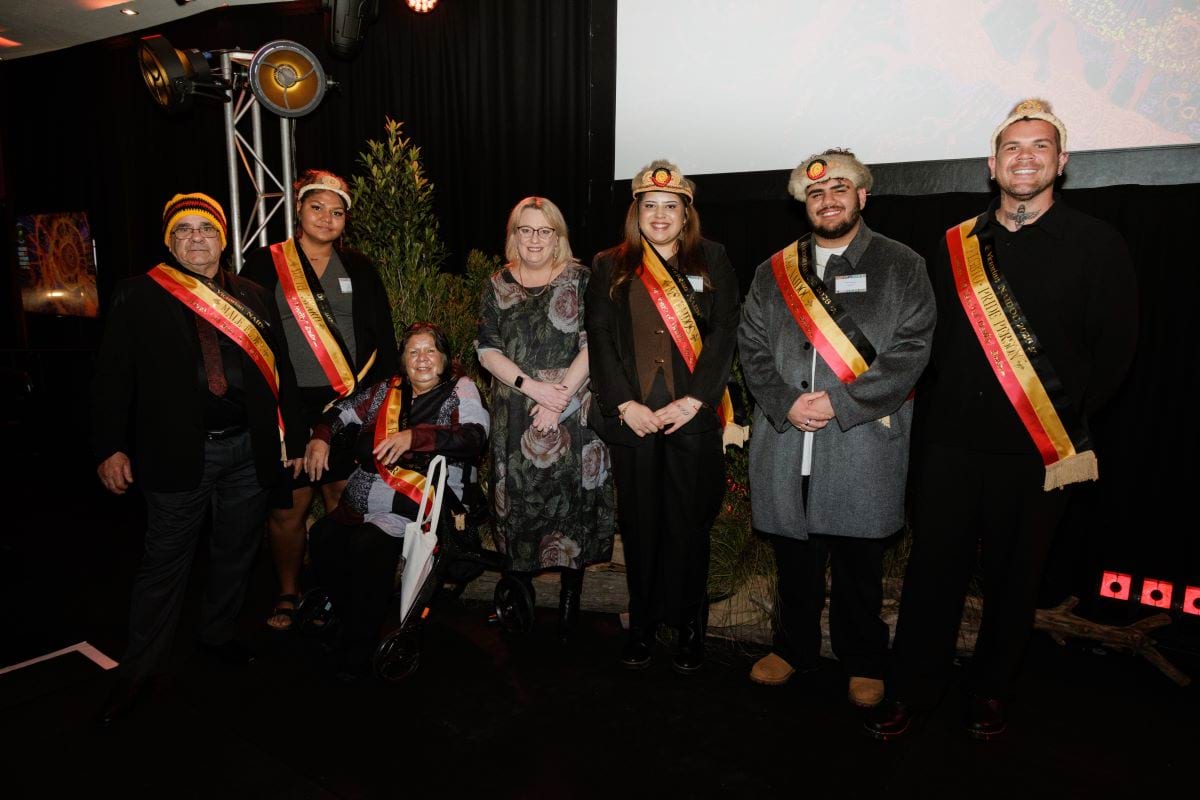 Photo of the Victorian NAIDOC Award Winners, Uncle Ross Morgan, Liana Liddle, Aunty Lyn Austin, Minister Ros Spence, Alkira Clark-Kickett, Adam Krakouer and Bebe Oliver. Standing on stage in a row wearing NAIDOC sashes and crowns.